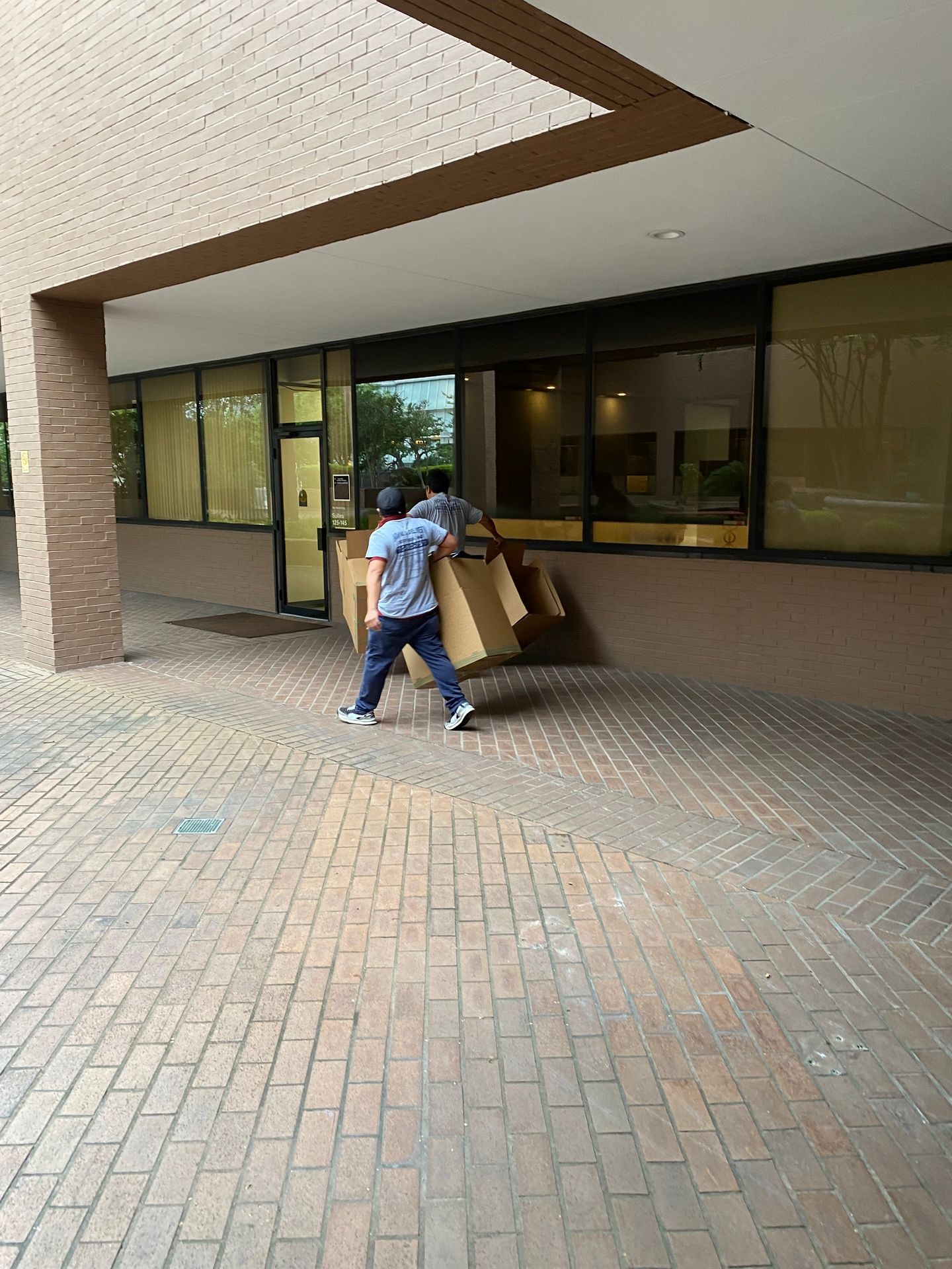 Two people carrying large cardboard boxes into a building with a brick exterior and large glass windows.