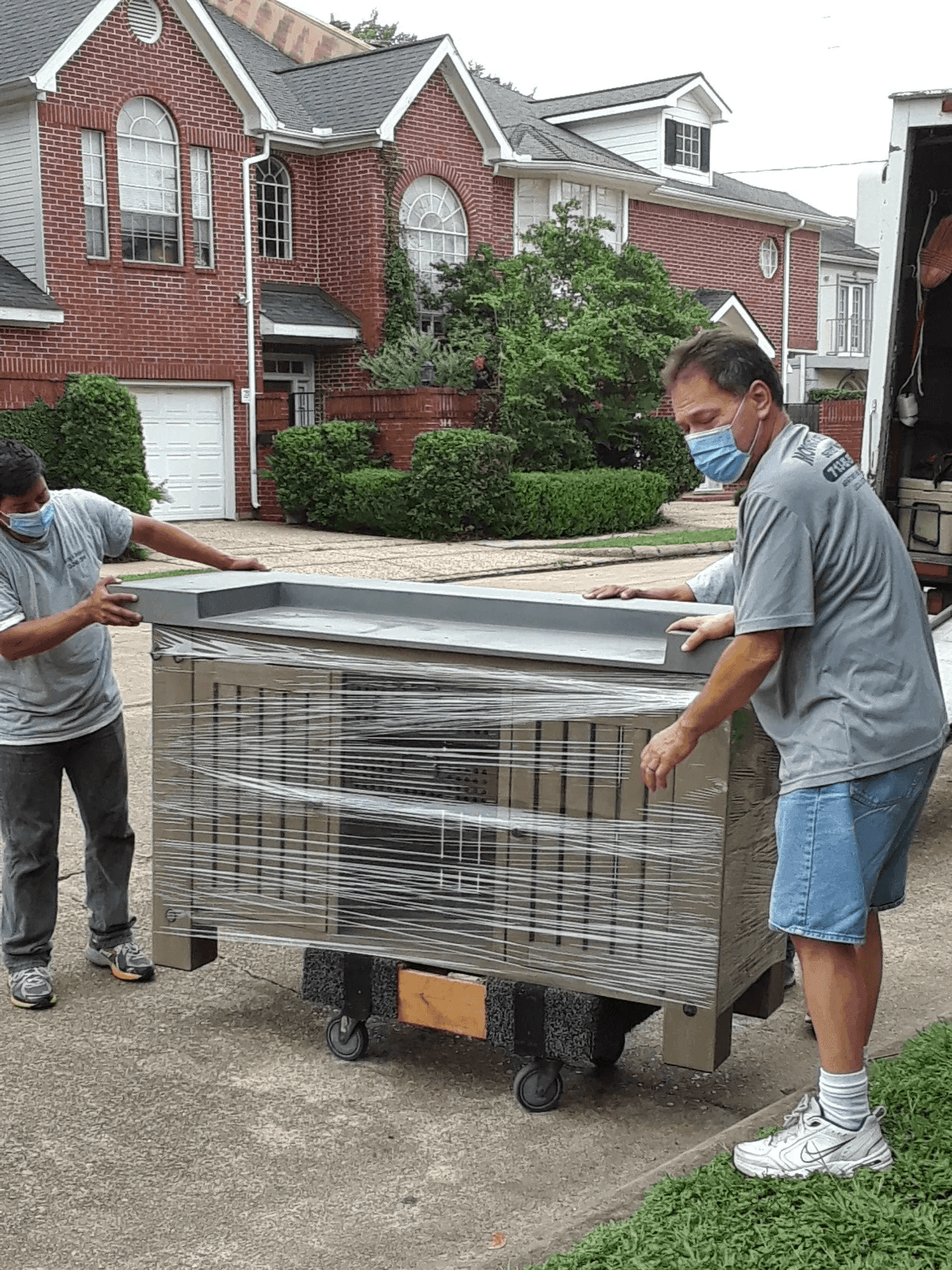 Two men wearing face masks moving large wrapped equipment on a dolly in front of brick houses.