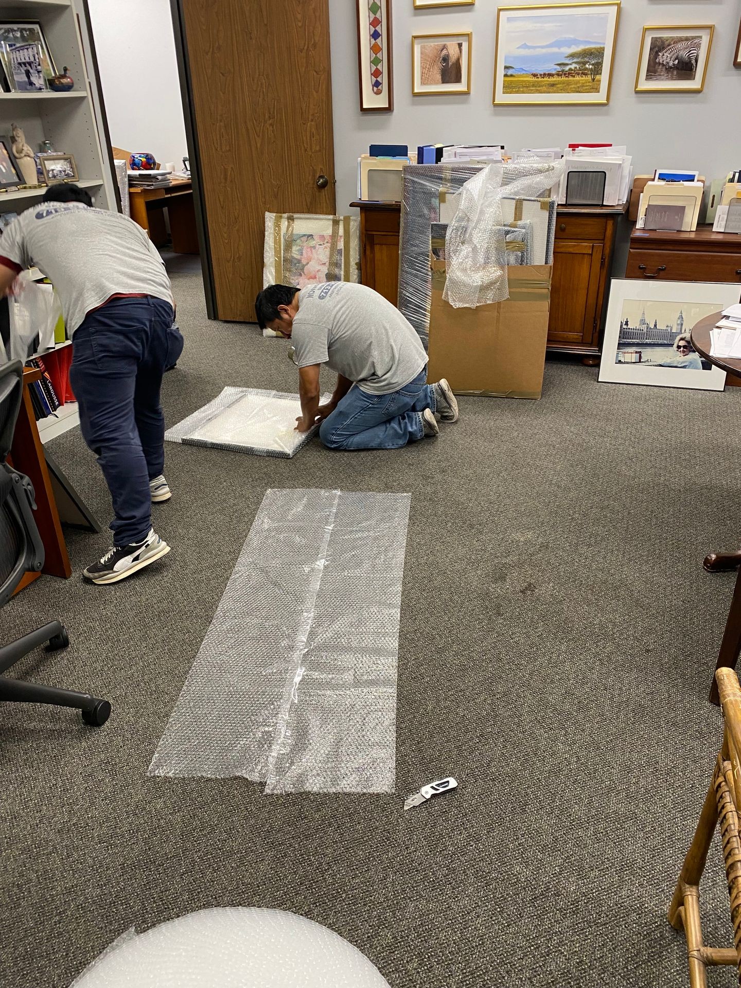 Two people wrapping framed pictures in bubble wrap on an office floor, surrounded by art and furniture.