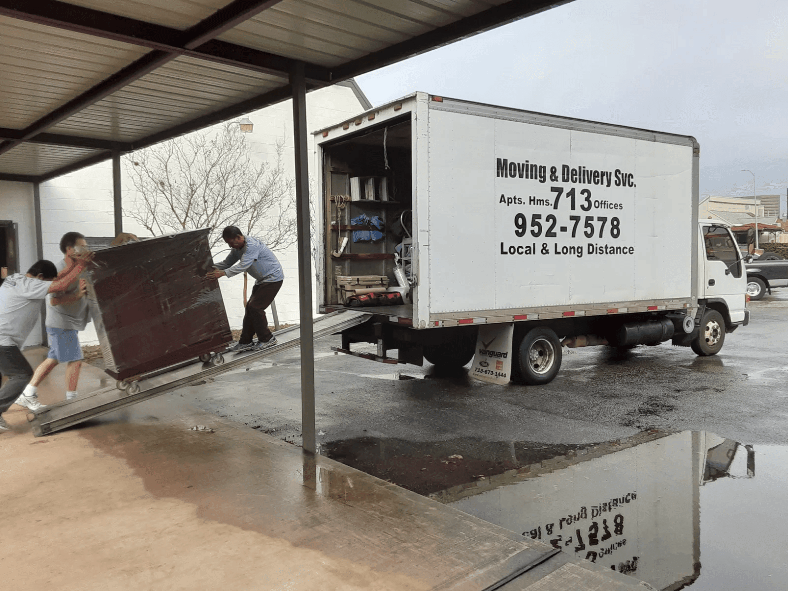 Movers loading a large item onto a truck ramp outside a building on a wet day.