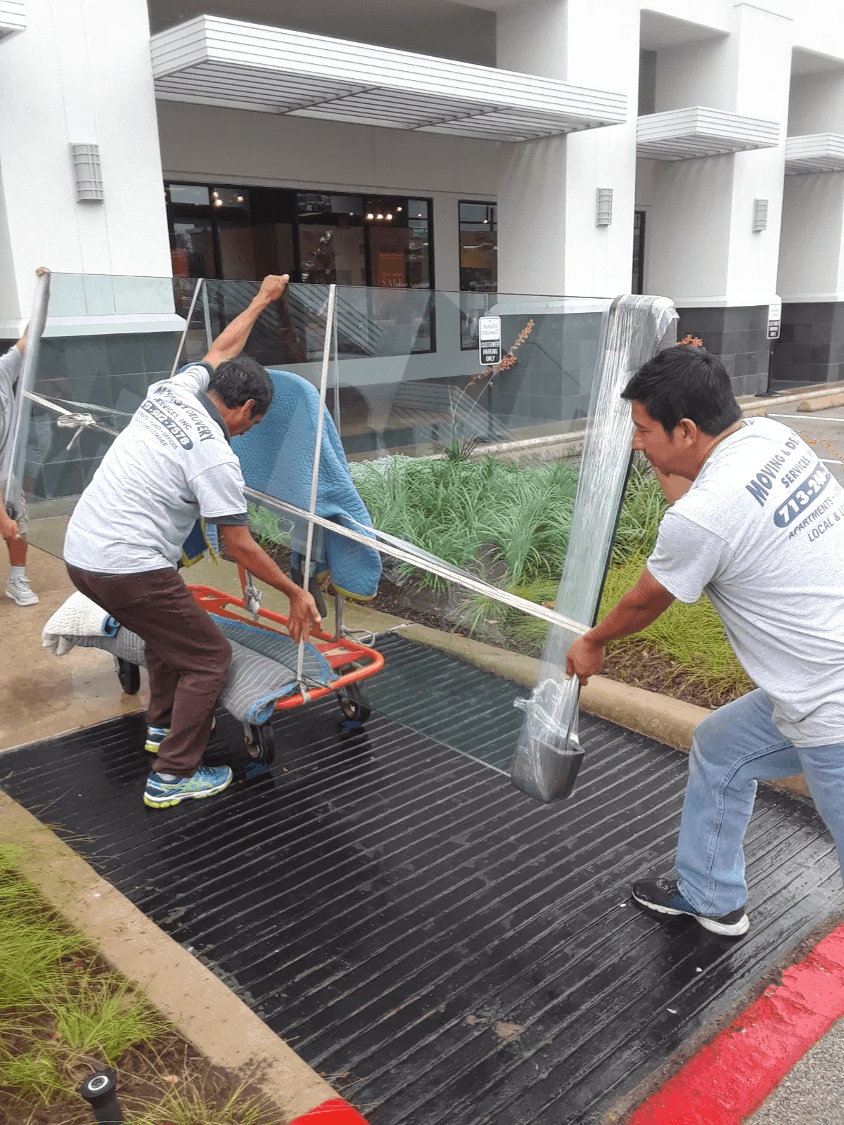 Two men carefully handling and transporting a large glass pane using moving equipment outside a building.